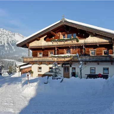 Ein traditionelles Holzhaus im Schnee mit Bergen im Hintergrund. Es ist ein klarer, sonniger Tag.