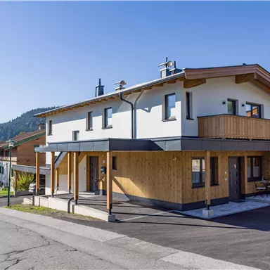 Ein modernes Haus mit Holzverkleidung und einem Balkon. Die Umgebung ist grün und von Bergen umgeben.