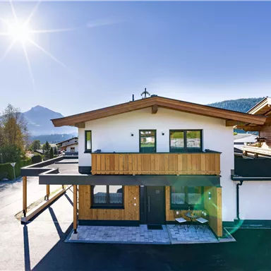 Ein modernes Haus mit Holzverkleidung und großen Fenstern. Im Hintergrund sind Berge und blauer Himmel sichtbar.