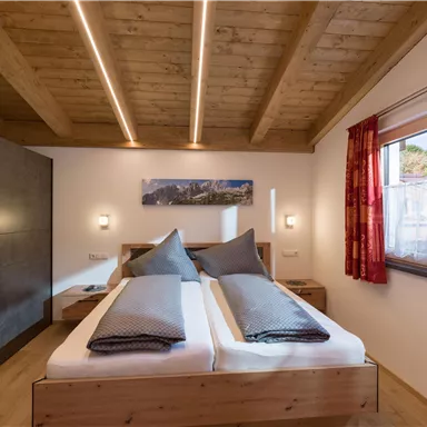 Ein modernes Schlafzimmer mit einem Doppelbett und Holzdecke. Große Fenster sorgen für viel Tageslicht und eine gemütliche Atmosphäre.