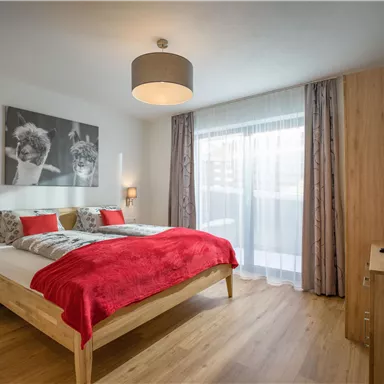 Ein gemütliches Schlafzimmer mit einem großen Bett und roten Dekokissen. Das Zimmer hat einen Holzfußboden und große Fenster, die viel Licht hereinlassen.