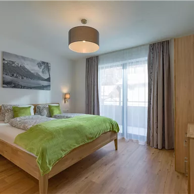 Ein modernes Schlafzimmer mit einem großen Bett und grüner Bettwäsche. Die Wände sind hell, und ein großer Fenster öffnet sich auf einen Balkon.