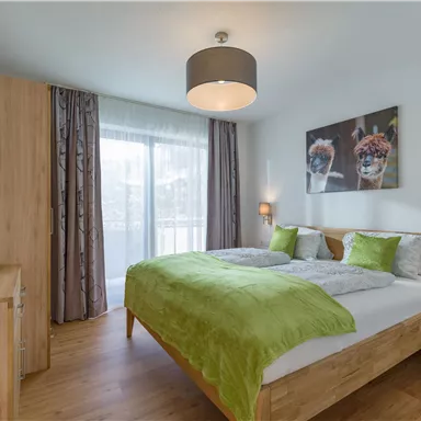 Ein modernes Schlafzimmer mit einem Doppelbett und hellgrüner Bettwäsche. Große Fenster sorgen für viel Tageslicht und eine angenehme Atmosphäre.