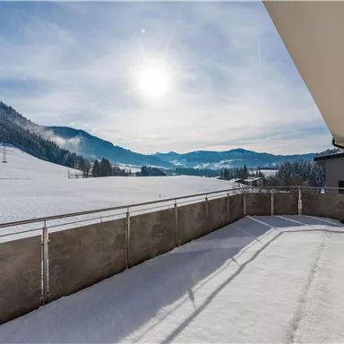 Eine winterliche Landschaft mit schneebedeckten Feldern und Bergen im Hintergrund. Die Sonne scheint hell am Himmel.