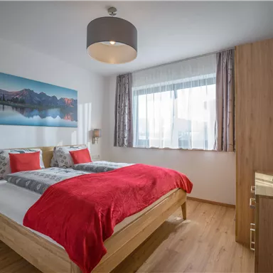 Ein modernes Schlafzimmer mit einem großen Bett und roten Kissen. Die Wände sind hell, und es gibt ein Fenster mit Vorhängen, das für Tageslicht sorgt.