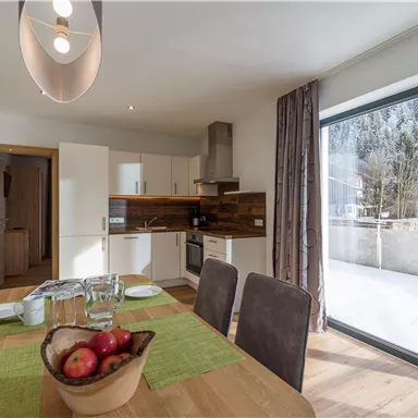 Eine moderne Küche mit einem Essbereich und einem großen Fenster, das einen Blick auf die Natur bietet. Der Tisch ist mit einer Schüssel Äpfel und Geschirr gedeckt.