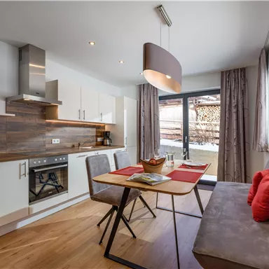 Eine moderne Küche mit Holzregalen und einer gemütlichen Essecke. Große Fenster sorgen für viel Tageslicht und einen Blick nach draußen.