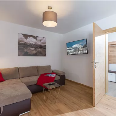 Ein gemütliches Wohnzimmer mit einer grauen Couch, roten Kissen und einem Fernseher an der Wand. Eine offene Tür führt zu einem weiteren Raum mit Holzdetails.