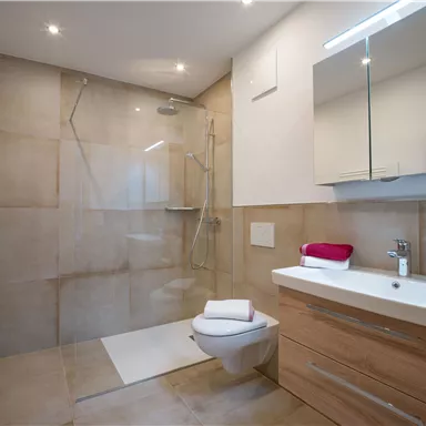 Ein modernes Badezimmer mit einer Glasdusche und einem Waschbecken aus Holz. Die Wände sind in neutralen Farben gehalten und es liegen zwei Handtücher bereit.