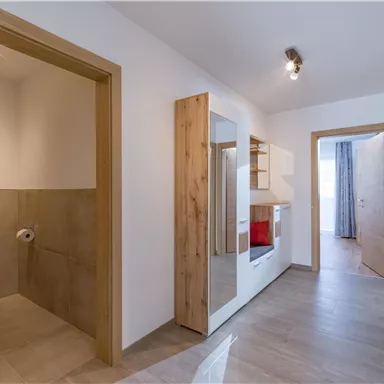 Ein moderner Flur mit hellen Wänden und einem Holztürrahmen. Auf der linken Seite ist ein Badezimmer und auf der rechten Seite ein Wohnbereich sichtbar.