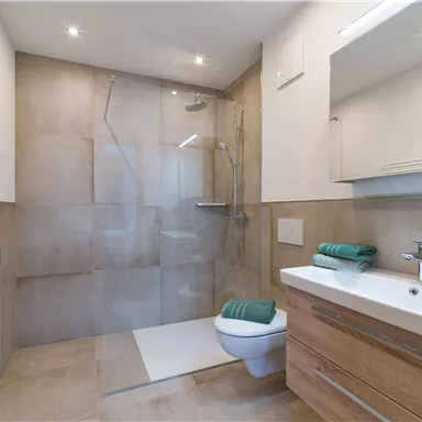 Ein modernes Badezimmer mit einer gläsernen Dusche und naturfarbenen Fliesen. Auf dem Waschtisch liegen grüne Handtücher und es gibt einen großen Spiegel.