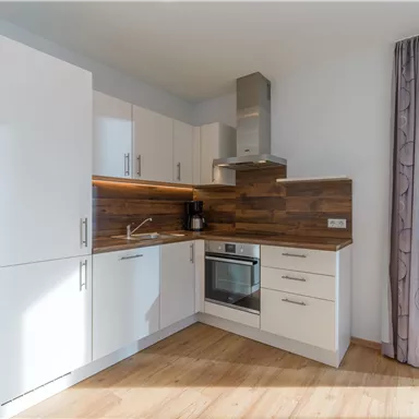 Eine moderne Küche mit weißen Schränken und Holzfronten. Sie verfügt über einen Backofen, ein Kochfeld und Fenster, die viel Licht hereinlassen.