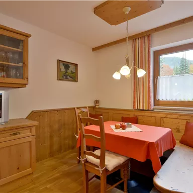 Ein gemütlicher Essbereich mit einem Holztisch und Stühlen. Die Wände sind hell und es gibt große Fenster, die Tageslicht hereinlassen.