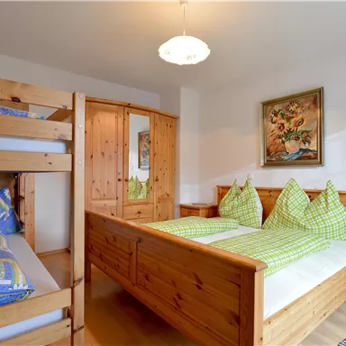 Ein gemütliches Schlafzimmer mit einem Doppelbett und einem Etagenbett. Die Wände sind aus hellem Holz und es gibt bunte Kissen und eine dekorative Wandmalerei.