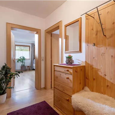 Ein heller Eingangsbereich mit Holzschränken und einem Spiegel. Im Hintergrund sind Pflanzen und ein weiterer Raum sichtbar.