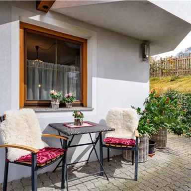 Ein gemütlicher Außenbereich mit einem kleinen Tisch und zwei Stühlen. Umgeben von Pflanzen und einer hellen, einladenden Atmosphäre.