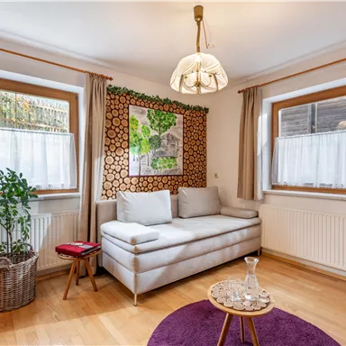 Ein gemütliches Wohnzimmer mit einer hellen Couch und einem runden Tisch auf einem lila Teppich. Das Zimmer hat große Fenster, die viel Tageslicht einlassen, und eine dekorative Wand mit Pflanzenmotiven.