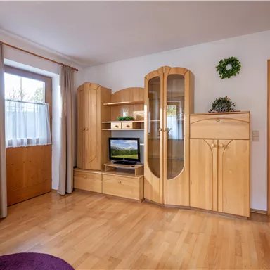 Ein gemütliches Wohnzimmer mit Holzmöbeln und einem Fernseher. Die Fenster bieten viel Tageslicht und der Boden ist aus Holz.