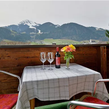 Ein gemütlicher Balkon mit zwei Stühlen und einem Tisch. Auf dem Tisch stehen Weingläser und eine Vase mit Blumen, während im Hintergrund die Berge zu sehen sind.