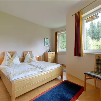 A cozy bedroom with two single beds, a chair, and windows that offer a view of the greenery. The room is bright and cheerfully designed.