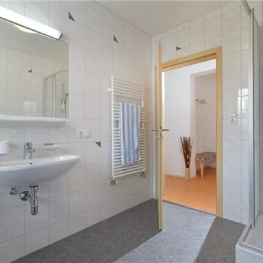 A modern bathroom with white tiles and a sink. In the background, a shower and access to another room are visible.