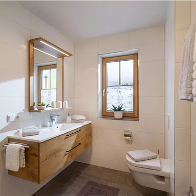 A modern bathroom with a wooden dresser and a large mirror. The window lets in natural light and there are fresh plants on the sink.
