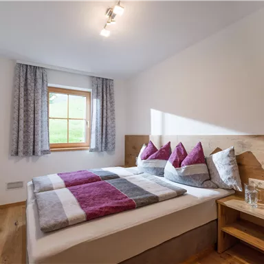 A cozy bedroom with a double bed and colorful pillows. Large windows and bright light create an inviting atmosphere.