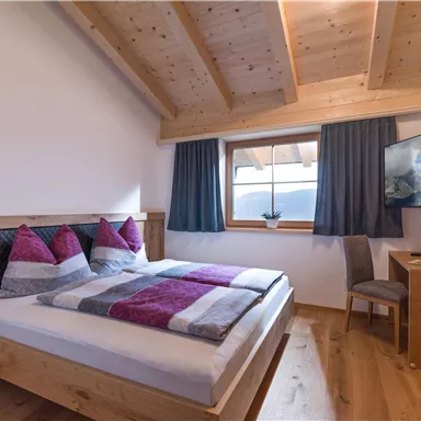 A cozy bedroom with a double bed and colorful cushions. The room has wooden paneling and a window with a view to the outside.