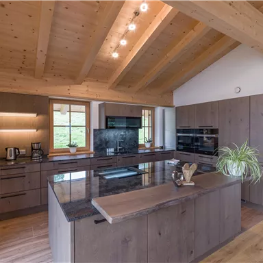 A modern kitchen with wooden cladding and large windows. The room is bright and welcoming, featuring a central kitchen island and modern appliances.