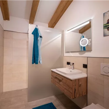 A modern bathroom with wooden ceilings and a bright tiled floor. It is equipped with a washbasin, a mirror, and color-coordinated towels.
