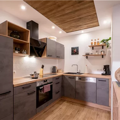 A modern kitchen with wood and gray elements. The equipment includes a stove, a sink, and open shelves.