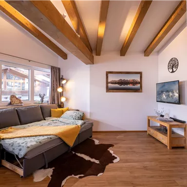 A modern living room with a gray sofa, wooden ceiling beams, and large windows. A television and a framed picture hang on the wall.