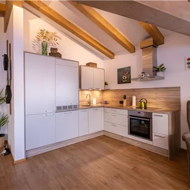 A modern kitchen with open shelves and wooden beams on the ceiling. The space is bright and inviting, featuring plants and contemporary furnishings.