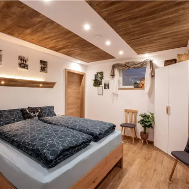 A cozy bedroom with a large bed and wooden ceiling. Pictures hang on the walls, and there is a plant as well as a chair in the room.