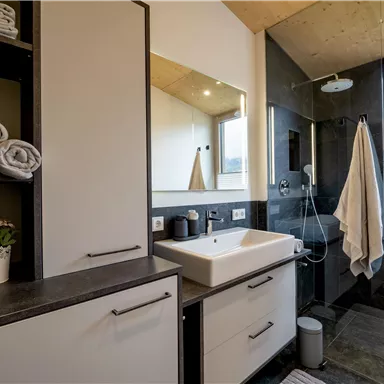 A modern bathroom with dark tiles and a bright sink. The shelves are decorated with fresh towels and a plant.