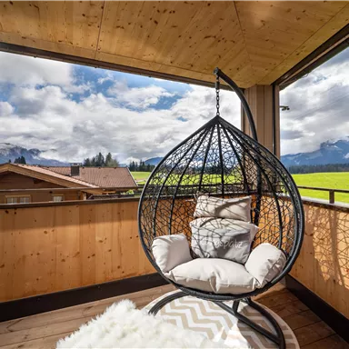 A cozy rocking chair on a terrace overlooking green meadows and mountains. The sky is partly cloudy, creating a relaxing atmosphere.