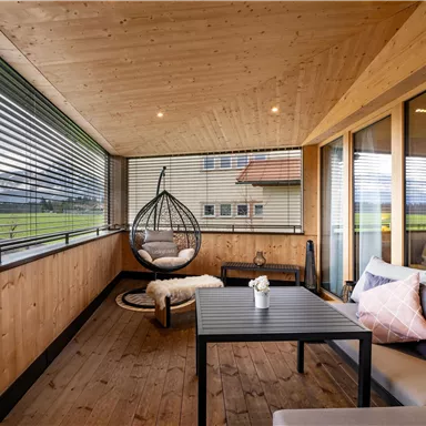 A cozy porch with wooden paneling and large windows. A hanging chair, a table, and comfortable cushions create a relaxing atmosphere.