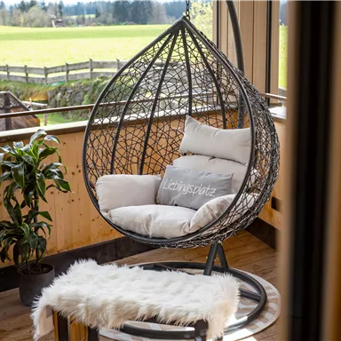 A cozy hanging chair made of rattan is located in a room with a large window overlooking the meadows. Next to it is a green plant and a small table with a soft rug.