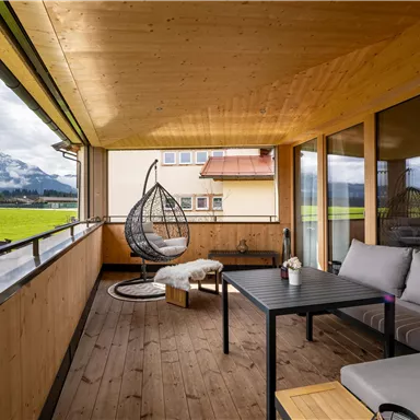 A cozy balcony with wooden cladding, a rocking chair, and views of green meadows and mountains. The seating area invites relaxation.