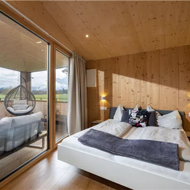 A cozy bedroom with wooden walls and a large bed. Through the windows, there is access to a balcony with a view of the landscape.
