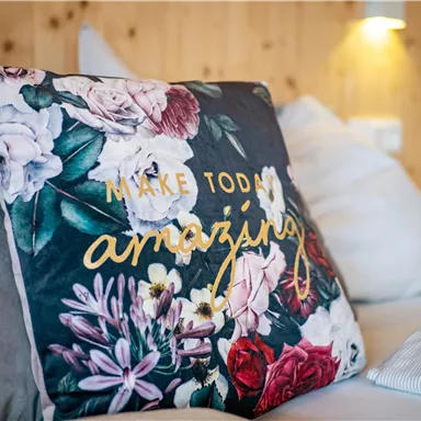 A decorated cushion with a floral pattern and the inscription "Make Today Amazing". It is placed on a sofa in a bright, cozy environment.