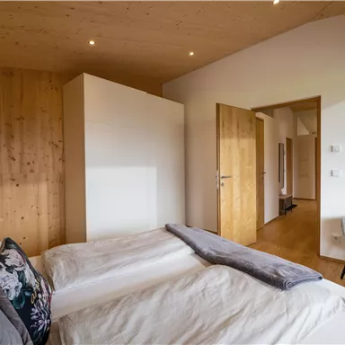 A modern bedroom with wooden paneling and a large window. The room is bright and cozy furnished, with a double bed and a chair.