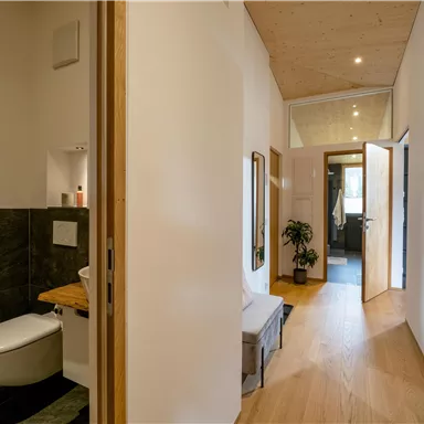 A modern hallway with an open door and wooden floor. On the left side is a small bathroom with elegant accents.