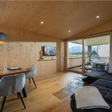 A modern living room with wooden interior and a large window. There is a dining table and a cozy seating area.
