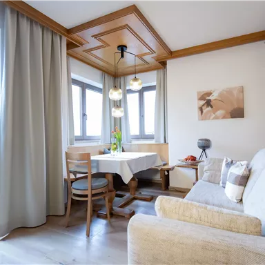 A cozy living area with a table, chairs, and a bright couch. Large windows provide plenty of daylight and a pleasant atmosphere.