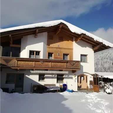 Ein schönes Holzhaus im Schnee mit einem Balkon und einer hellen Fassade. Der Himmel ist blau und der Bereich ist von einer ruhigen Winterlandschaft umgeben.