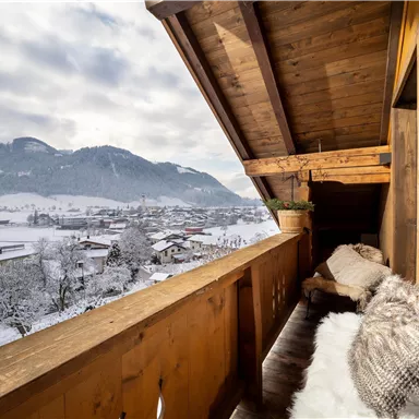 A cozy balcony with wooden cladding overlooks a snowy landscape. In the background, mountains and houses are visible.