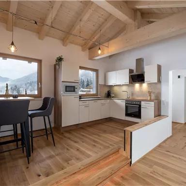 A modern kitchen with light wooden furniture and large windows. The room has a cozy atmosphere and a view of the mountains.