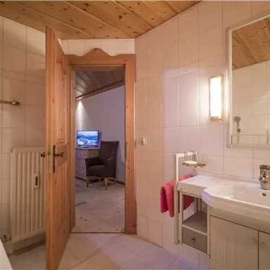 Ein modernes Badezimmer mit hellen Fliesen und einem Waschbecken. Durch die offene Tür ist ein gemütlicher Wohnbereich mit Fernseher sichtbar.