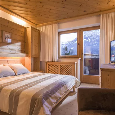 Ein gemütliches Schlafzimmer mit holzverkleideten Wänden und einem großen Fenster, das einen Blick auf die Berge bietet. Das Zimmer ist modern und einladend eingerichtet, mit einem Fernseher und einem komfortablen Sessel.